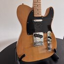 Elektrická kytara typu Telecaster v přírodní dřevěné barvě s černým pickguardem. Nástroj je umístěn na černém stojanu a nemá žádné viditelné značky a…