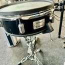 Černý buben s chromovaným obručem a ladidly, umístěný na kovovém stojanu. Na korpusu je bílý štítek s nápisy „Roland“, „Acoustic Design“ a „V-Drums“.…