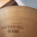 Detailní záběr na povrch mosazného činele. Na povrchu je vyryt nápis TOP NEW BEAT HI HAT. Povrch vykazuje koncentrické drážky typické pro opracování…