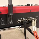 Detailní pohled na zadní panel červeného klávesového nástroje s černým spodním rámem. Jsou vidět různé konektory a popisky: AM UP/DN PEDAL, USB, ROTO…