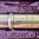 Detail stříbrného kovového dílu flétny v fialovém polstrování pouzdra. Na povrchu je vyryto TAKUMI, NISHIMA FLUTE COLL. a JAPAN. Povrch vykazuje drob…
