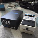 Bílá efekt pedál s nápisy Digital Delay a DD-8 a logem BOSS. Na horní části jsou čtyři modré ovládací knoflíky a konektory s popisky OUTPUT A, OUTPUT…