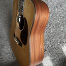 Akustická kytara s přirozeným dřevěným povrchem a tmavou lemovkou. Tělo má hnědý odstín s viditnou strukturou dřeva a tmavý pickguard s mramorovým vz…