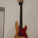 Světle hnědá elektrická basová kytara s přírodním dřevěným povrchem a červeným pickguardem. Na hlavě hmatníku je nápis Fender PRECISION. Nástroj má z…