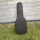 Černý poloměký obal (gig bag) na kytaru s dlouhým hnedkem. Obal je vyroben z textilního materiálu a vybaven dvěma popruhy s plastovými přezkami. Povr…