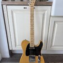 Elektrická kytara s přírodním dřevěným povrchem těla a hmatníkem. Na hlavě je viditelné logo Fender. Kytara má černý pickguard a chromované potenciom…