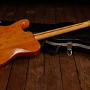 Elektrická kytara s přírodním dřevěným povrchem a světlým hnedým hmatníkem leží na černém tvrdém kufru. Kytara má tvar typu Telecaster a je zobrazená…