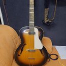 Polodupatá elektrická kytara s korpusem v přechodové barvě od žluté po černou. Nástroj má bílý plechový kryt (pickguard) a bílé ladičky na hlavě. Na…