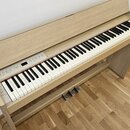 Digitální piano v dřevěném korpusu světlé barvy s bílými a černými klávesami. Na ovládacím panelu jsou umístěny tlačítka a nápisy, včetně označení "G…