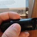 Detailní pohled na černý, lesklý plastový předmět cylindrického tvaru, který drží ruka. Povrch vykazuje drobné škrábance a prachové částice. V pozadí…