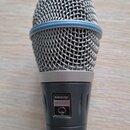 Dynamický mikrofon s kovovou síťkou a modrým prstenem v horní části. Na těle je viditelný nápis SHURE a model BETA 87A. Povrch mikrofonu je černý, v…