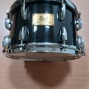Černý buben s chromovaným obručováním a ladidly. Na korpusu je zlatý štítek s nápisy Pearl, MAPLE SHELL, 000170 a MADE IN JAPAN. Povrch nástroje je g…