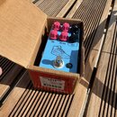 Modře barevný efekt pedál v kartonové krabici. Na pedálu jsou čtyři červené a dva černé oválné knofíky s popisky TONE, MID FREQ a PUSH. Uprostřed je…