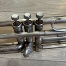Detailní pohled na stříbrnou trubku s třemi ventily. Na horních krytkách ventilů jsou bílé perleťové vložky a pod nimi jsou vidět červené plsti. Povr…