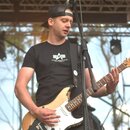 Muž v černém tričku a kšiltovce hraje na elektrickou kytaru typu Stratocaster. Kytara má přírodní dřevěnou barvu s výraznými odřenými černými plocham…