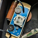 Modrá tiskovaná spoje s elektronickými komponenty, dvěma černými otočnými potenciometry a kovovým konektorem. Na desce je svisle napsaný bílý text 11…