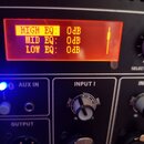 Detailní záběr na ovládací panel elektronického zařízení s červeným digitálním displejem. Na displeji je text HIGH EQ: 0dB, MID EQ: 0dB a LOW EQ: 0dB…