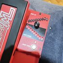 Červený kontrolér s černým ovládacím panelem a stříbrným knoflíkem. Na panelu jsou nápisy SHIFT UP, SHIFT DOWN a DROP TUNE, řady čísel 1 až 8 s označ…