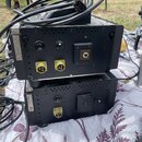 Dvě černé kostky elektronických zařízení s perforovaným horním krytem, umístěné na sobě. Jsou vidět různé konektory, včetně dvou žlutých vícepinových…