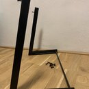 Černý kovový stojan v rozloženém stavu, který stojí na dřevěné podlaze. Na horních hranách dvou svislých částí jsou vyčnívající šrouby a na zemi leží…