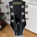 Záběr na hlavu elektrické kytary v černé barvě s bílým lemováním. V horní části je zlatý nápis GRETSCH. Jsou vidět šest chromovaných ladiček a struny…
