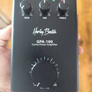 Černý kytarový výkonový zesilovač s nápisy Harley Benton a GPA-100 Guitar Power Amplifier. V horní části jsou tři ovládací knoflíky s popisky BASS, M…