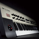 Stříbročerný syntetizátor s klávesnicí a ovládacím panelem. Na horní části je nápis KORG Kronos SYNTHESIZER. Panel obsahuje řadu černých otočných ovl…