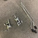 Na šedém koberci leží dvě chromované mechanismy pedálů a dvě kovové tyče s černými gumovými koncovkami. Na kovových částech nejsou vidět žádné značky…