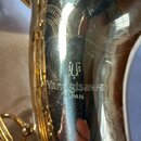 Detailní záběr na zlatý, lesklý povrch dechy saxofonu. Na povrchu je vyryto logo znázorňující stylizované písmeno U a nápisy Yanagisawa JAPAN. Povrch…