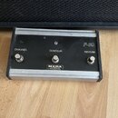 Černý ovládací panel s hliníkovým rámem, na kterém jsou tři přepínače. Na panelu jsou bílé nápisy CHANNEL, CONTOUR a REVERB, v pravém horním rohu ozn…