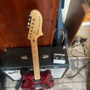 Elektrická kytara s tmavě červeným trupem, světlým hnedým krkem a hlavou. Na hlavě je nápis Squier STARCASTER. Kytara je opřena o černý a stříbrný ko…