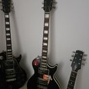 Tři černé elektrické kytary s tvarem typu Les Paul. Středová kytara má na hlavě nápis "Lijamani", na těle nálepky s texty "esp" a "ensa" a podpis. Le…