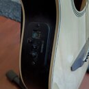 Detailní pohled na bok akustické kytary světlé barvy s černým ovládacím panelem. Na panelu je nápis YAMAHA, displej ladičky a ovladače s popisky TUNE…
