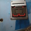 Detailní pohled na černý plastový konektor pro kabel, který je přišroubován k modrému povrchu nástroje. Konektor má červené vnitřní prvky a na horní…