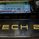 Modrý efekt pedál s nápisy TECH 21 a STEVE HARRIS je umístěn v černé kovové krabičce spolu s černým napájecím adaptérem. Na horní části pedálu je zob…