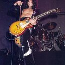 Fotografie zachycuje hudebníka s elektrickou kytarou typu Les Paul v žlutém goldtop provedení s červeným lem. Kytara má dva humbuckery a bílé ovládac…