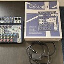 Na fotografii je kompaktní mixážní pult s modrým šasi a bílými ovládacími panely, krabice od produktu a černý kabel s konektorem. Na zařízení i na kr…