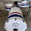 Bílá bicí souprava s červeným a modrým vodorovným pruhem. Na přední straně basového bubnu je černé logo s nápisy SINCE 2004, IDC DRUMS a QUALITY HAND…