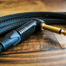 Černý audio kabel s pleteným povrchem a pozlaceným 6,3mm jack konektorem. Konektor má černý plastový úhelníkový kryt s žebrovaným detaillem. Na kabel…