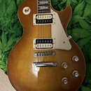 Elektrická kytara s trupem v hnědém goldtop odstínu a krčkem s hmatníkem z tmavého dřeva. Nástroj má dva humbuckery v krémových rámečkách, krémový pi…