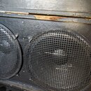 Detailní pohled na černý reproduktorový box s dvěma velkými membránami krytými kovovými mřížkami. Povrch boxu je z černého lakovaného dřeva s viditel…