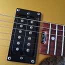 Detailní pohled na hmatník a snímač elektrické kytary v žluté barvě. Černý humbucker je osazen v černém rámečku a má viditelné šrouby. Hmatník je vyr…