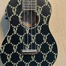 Černý ukulele s celoplošným zlatým vzorem stylizovaných lidských postav a malých čtverců. Uvnitř ozvučnice je štítek s nápisy „BILL WISH SIGNATURE UK…