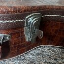 Detailní záběr na hnědý kufřík s povrchem imitujícím krokodýlí kůži a bílým stehem. Na kufříku je vidět kovový zámek s vloženým klíčem a část dalšího…
