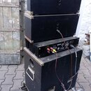 Stoh černých reproduktorových beden a jednoho zesilovače umístěný na vozíku. Zesilovač má na zadním panelu nápisy OUTPUT, SEAK MONO 100W/JET a nečite…