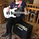 Žena drží bílou elektrickou kytaru značky Jackson s černým hmatníkem a černými snímači. Kytara je připojena k černému kombu s nápisem Marshall. Na ze…