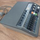 Šedý obdélníkový přístroj s nápisem KORG RHYTHM 250. Přední panel obsahuje řadu černých čtvercových tlačítek s bílými popisky rytmů, několik otočných…