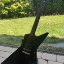 Černá elektrická kytara s ostře hranatým tělem typu Explorer umístěná na černém stojanu. Na hlavě hmatníku je bílý nápis Gibson. Kytara má dva humbuc…