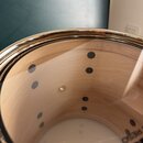 Detailní pohled do vnitřku světlého dřevěného korpusu bubnu. Na vnitřní stěně jsou vidět tmavé kulaté body a jeden světlý kovový šroub. V dolní části…