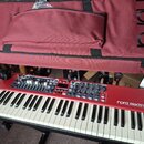 Červený digitální klavír s bílými a černými klávesami. Na pravé straně je bílý nápis nord electro 6D STAGE PIANO / KEYBOARD SYNTHESIZER. Nad klávesam…
